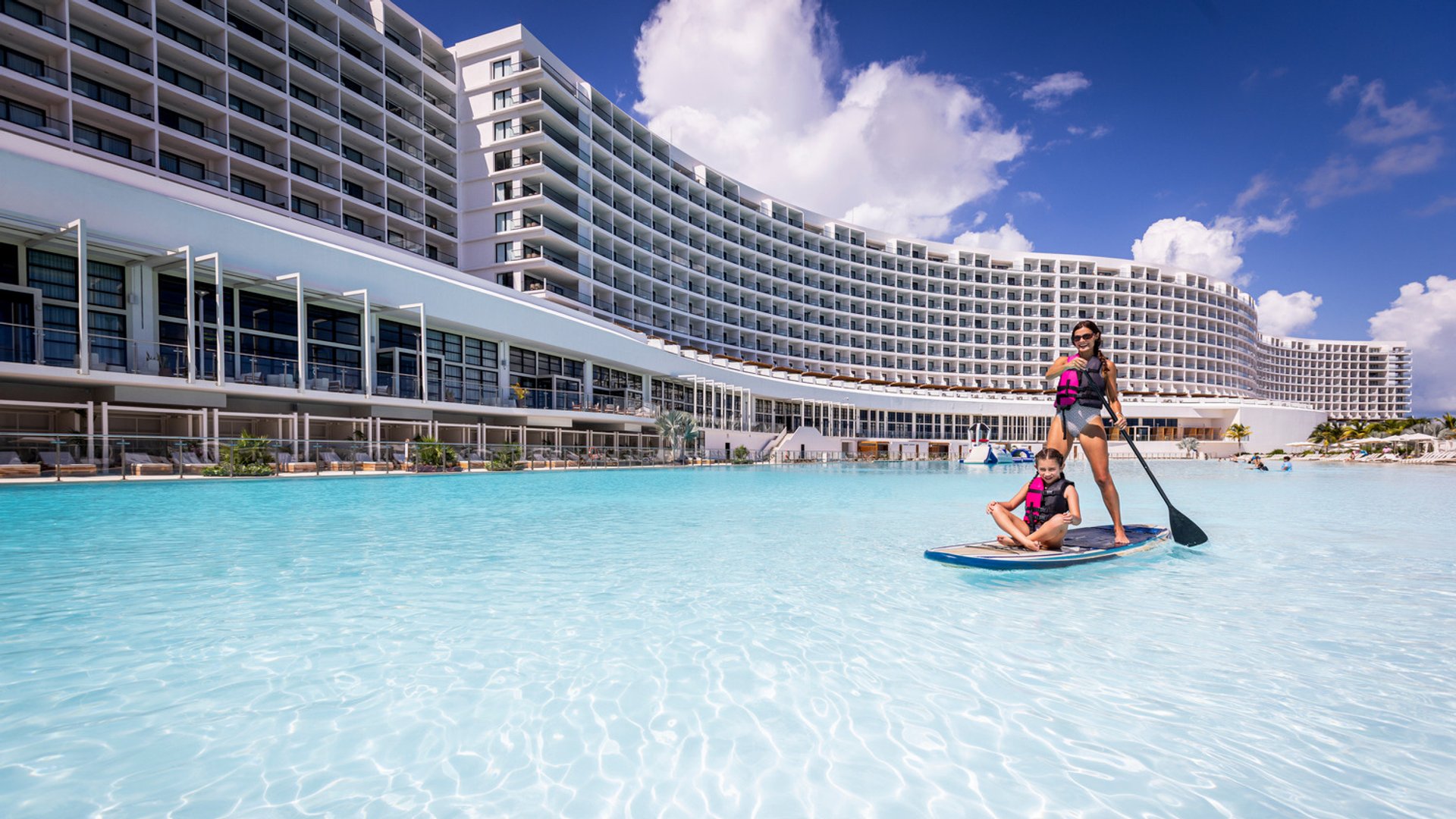 Two people on a paddleboard in a turquoise pool near a large, curved resort building under a clear blue sky.