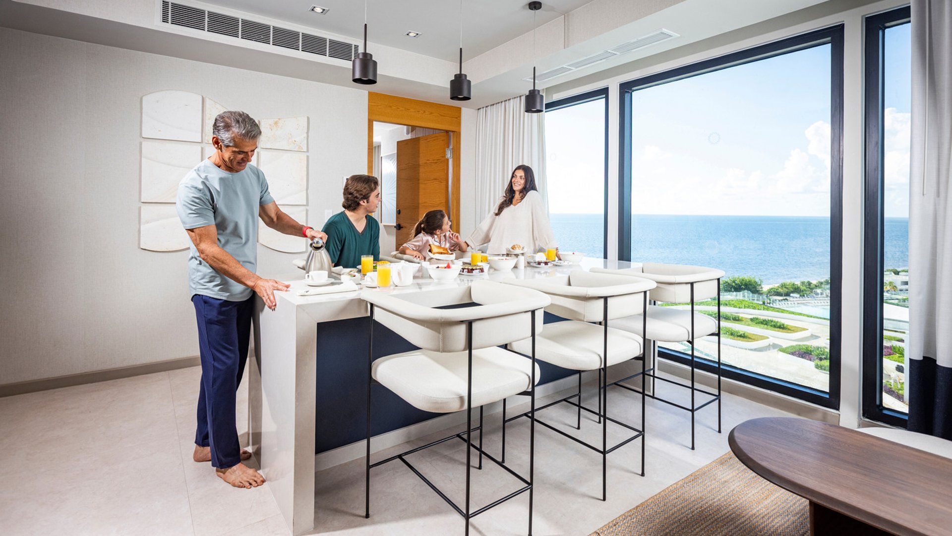 Family in pajamas dining in a room with a large ocean-view window, sunny day outside, modern interior design.